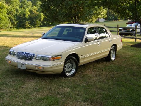 1996 Mercury Grand Marquis - A cream-colored sedan is parked on a grassy lawn with trees in the background and another vehicle behind a wooden fence.