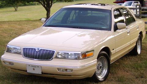 1996 Mercury Grand Marquis - A tan 1996 Mercury Grand Marquis is parked on a grassy lawn, showing its distinctive chrome grille and wire wheels.