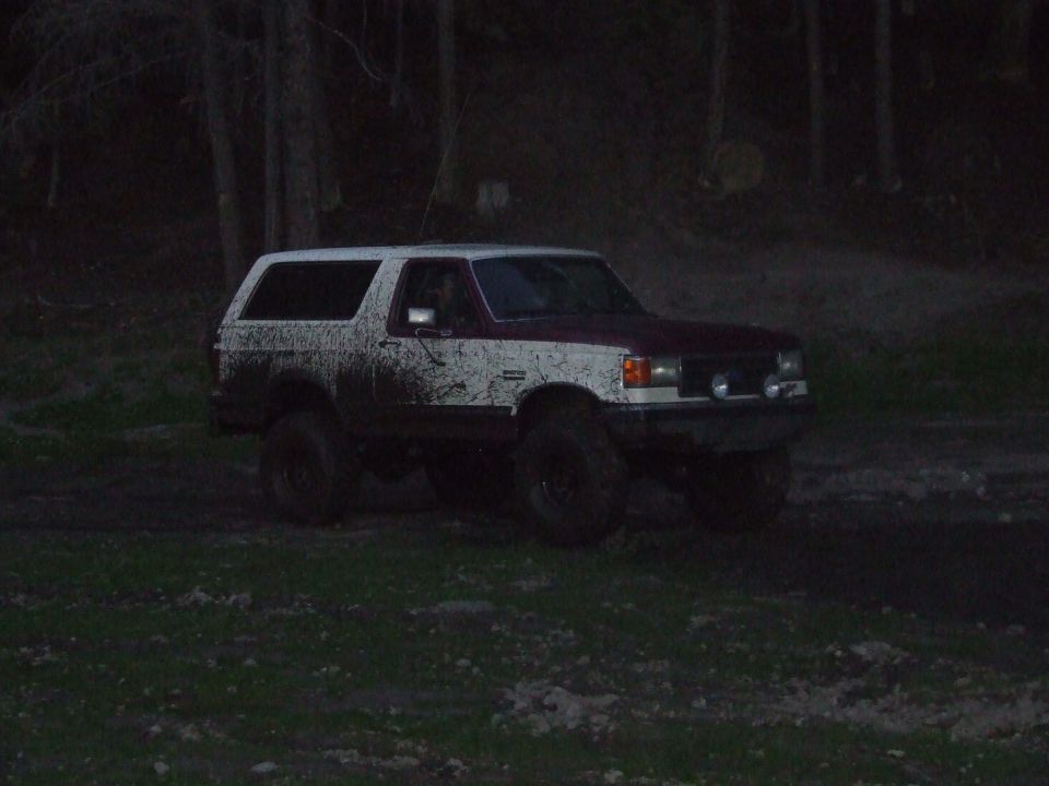 1990 Ford Bronco covered in mud, parked on grassy terrain with trees in background