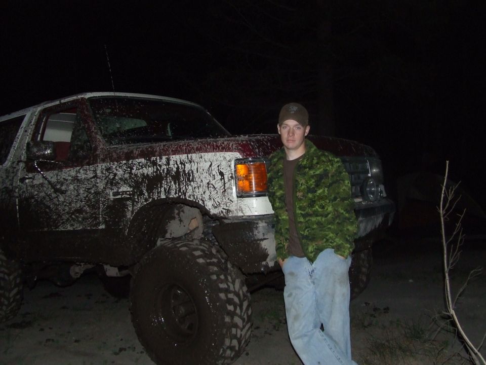 1990 Ford Bronco covered in mud, with a man in a camouflage jacket standing beside it at night