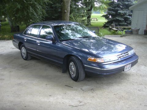 1996 Ford Crown Victoria - A blue sedan is parked outdoors on a dirt surface with trees and a building in the background.