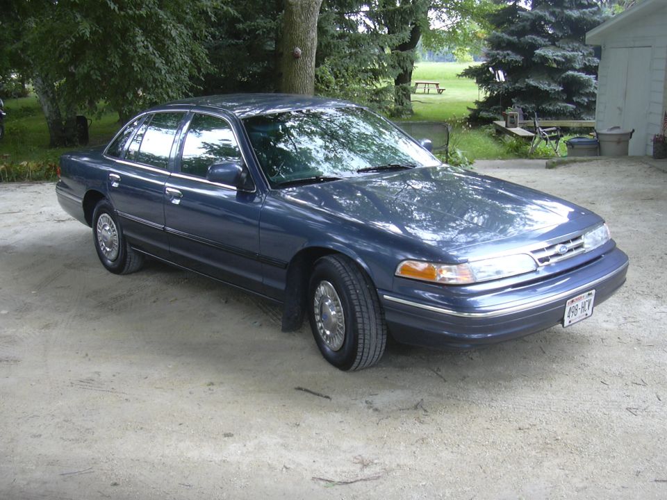 Blue sedan parked on dirt, front quarter view, chrome wheels, visible license plate