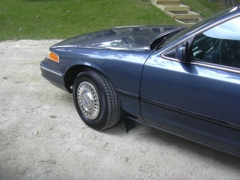 1996 Ford Crown Victoria - The front fender, wheel, and side profile of a blue 1996 Ford Crown Victoria are visible in this image.