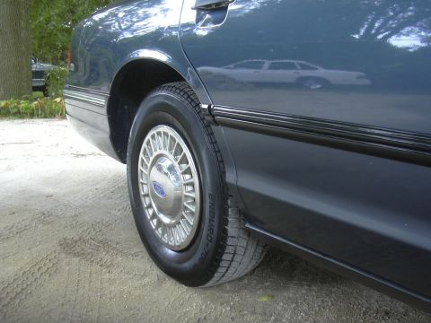 1996 Ford Crown Victoria - Rear side view of a dark blue 1996 Ford Crown Victoria, showing the wheel and quarter panel. Another car is visible in the reflection.