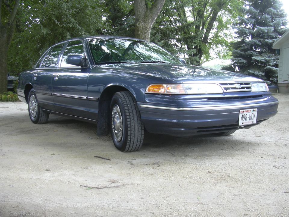Dark blue Ford Crown Victoria parked on a dirt lot with trees and a house in the background. License plate reads 498-HCV