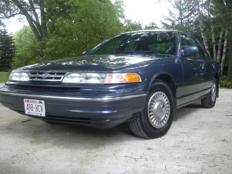1996 Ford Crown Victoria - A dark blue sedan is parked outdoors on a gravel surface with trees in the background.