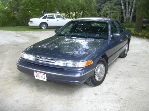 1996 Ford Crown Victoria - Two Ford Crown Victorias, one dark blue and one white, parked outdoors on a gravel surface.