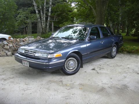 1996 Ford Crown Victoria - This sedan is parked outdoors with trees and a wooden structure in the background.