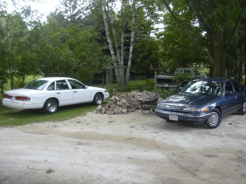 1996 Ford Crown Victoria - Two Ford Crown Victoria sedans, one white and one dark blue, are parked in a yard with trees and a pile of bricks.