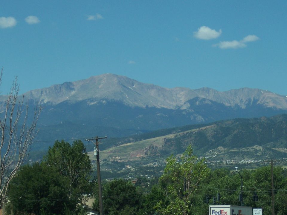 Mountains under a blue sky with scattered clouds. Trees and a utility pole are in the foreground. A FedEx truck is partially visible