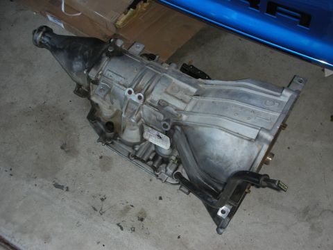 1998 Ford Crown Victoria - An automatic transmission is shown resting on a concrete floor, with a blue vehicle in the background.