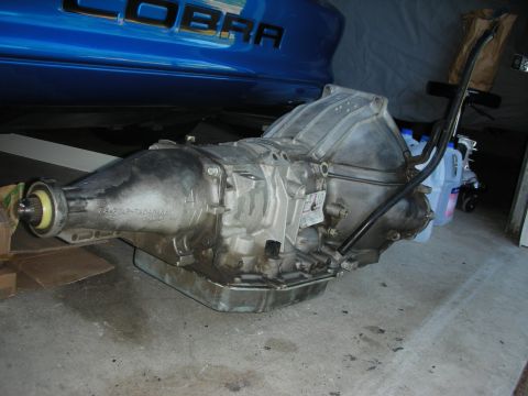 1998 Ford Crown Victoria - A used automatic transmission sits on a concrete floor, with part of a blue car bearing the word "COBRA" visible above it the background.