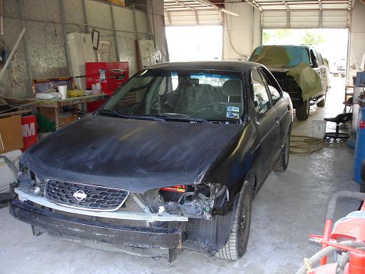 Nissan sedan with front bumper removed, in a garage with another vehicle covered in the background
