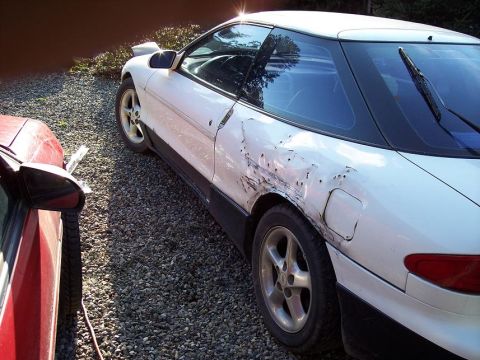 1993 Ford Probe - A white coupe with extensive damage to its side is parked outdoors next to another vehicle.