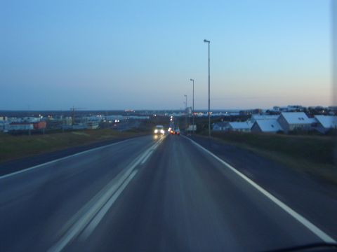 1974 Ford Bronco - Driving on a highway towards a town as the sky darkens, with streetlights and distant buildings visible.