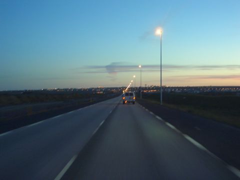 1974 Ford Bronco - A lone van travels down a highway as the sun sets, with city lights visible on the horizon.