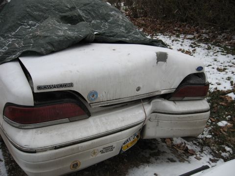 1992 Ford Crown Victoria - The rear of a white Ford Crown Victoria, partially covered by a tarp, shows damage to the trunk and bumper.
