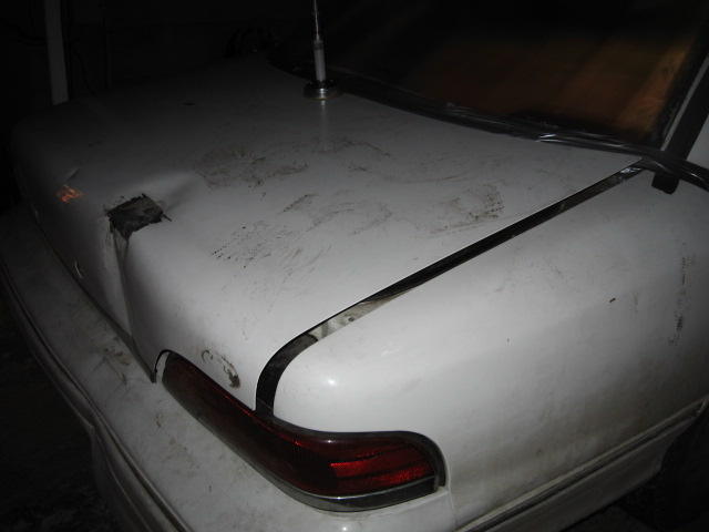 Rear of a white sedan with significant damage to the trunk lid and quarter panel. Visible dent and missing paint
