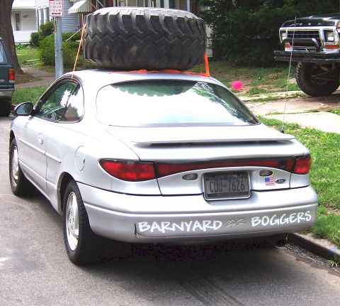 1978 Ford Bronco - A silver sedan is parked on a street with a large tire secured to its roof rack.