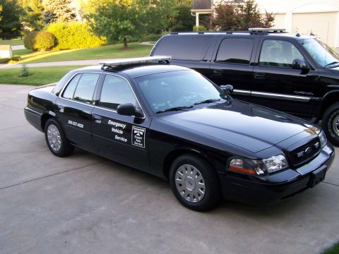 2003 Ford Crown Victoria - A black Ford Crown Victoria, marked as an emergency vehicle, is parked in a driveway next to a large black SUV.