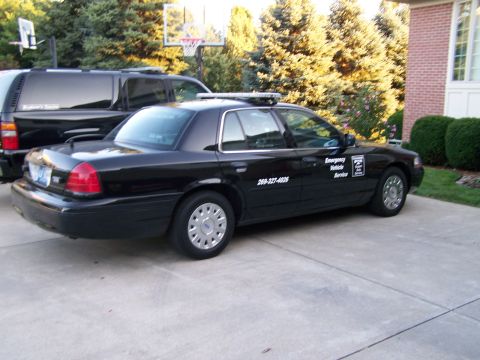 2003 Ford Crown Victoria - A black 2003 Ford Crown Victoria sedan, marked as an emergency vehicle, is parked in a driveway next to an SUV.