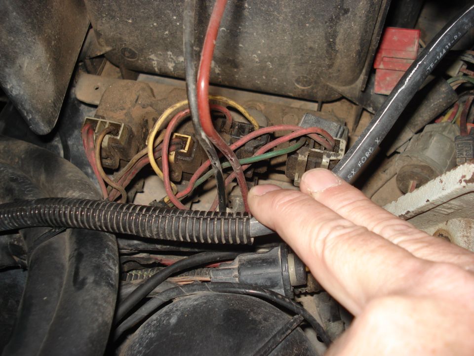 Close-up of automotive fuse box with red, yellow, and green wires. A hand is pointing to a connection