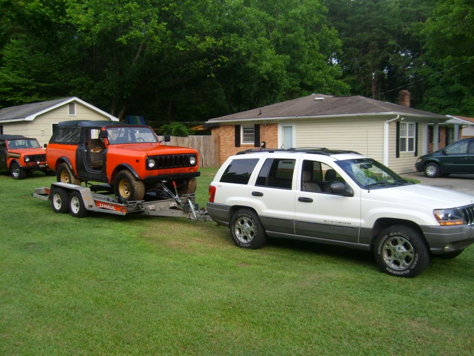 White Jeep Grand Cherokee towing an orange International Scout on a U-Haul trailer, with another orange vehicle visible behind the Scout