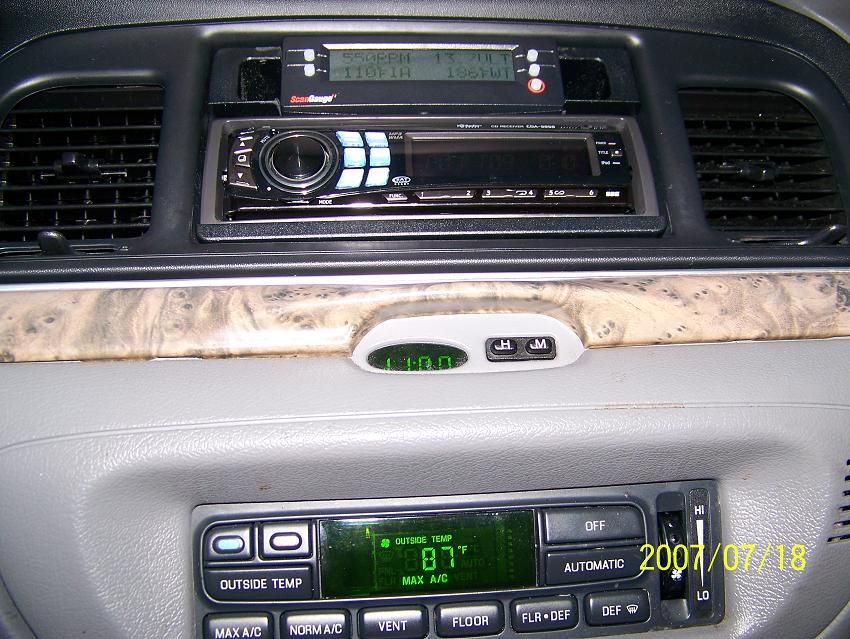 Car dashboard showing two stereos stacked, air vents, and climate controls. A digital clock is visible
