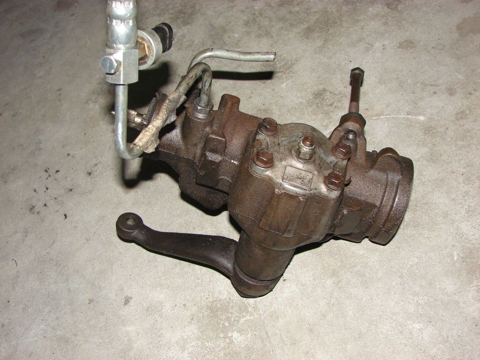 Rusty steering gear box with hydraulic lines attached, resting on a concrete surface