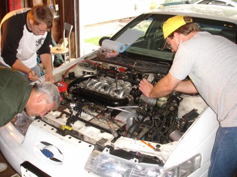 1993 Ford Taurus - Three men are gathered around the open engine bay of a white 1993 Ford Taurus, appearing to work on the engine.