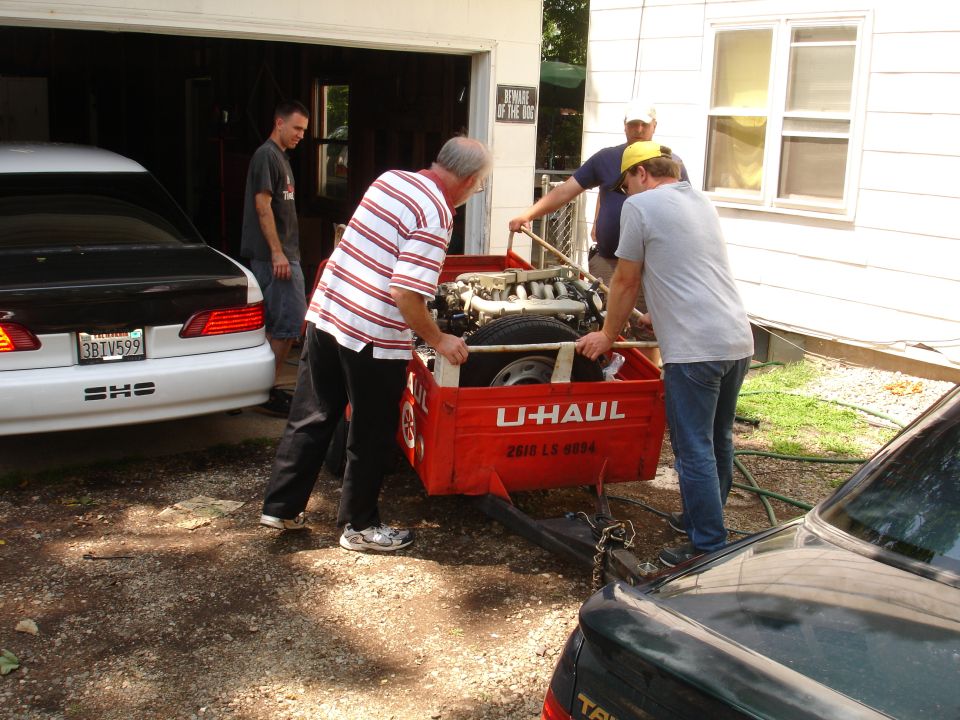 Four guys are loading a V8 engine into a U-Haul trailer next to a white car