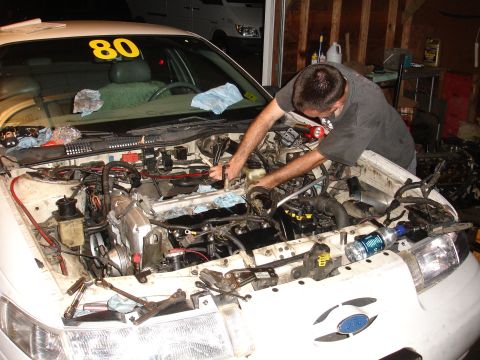 1993 Ford Taurus - Mechanic works on the engine of a white car, with a yellow number 80 visible on the windshield.