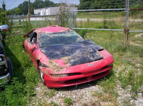 1995 Ford Probe - This damaged red coupe, believed to be a 1995 Ford Probe, sits abandoned in a grassy area.