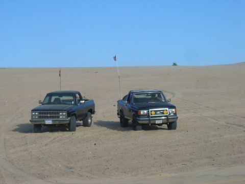 1989 Ford Bronco - Two pickup trucks are parked on a sandy dune under a clear blue sky with flags on poles.