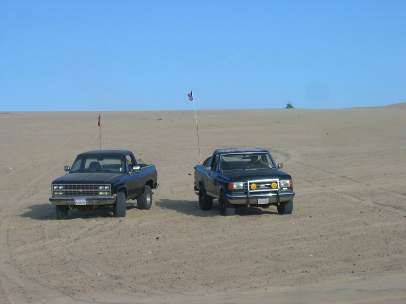 Two pickup trucks are parked on a sandy dune under a clear blue sky. One truck is black, the other is dark blue