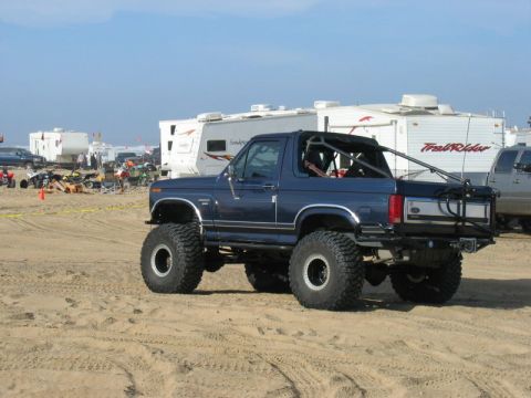 1989 Ford Bronco - Modified blue Ford Bronco parked on sand with RVs in the background.