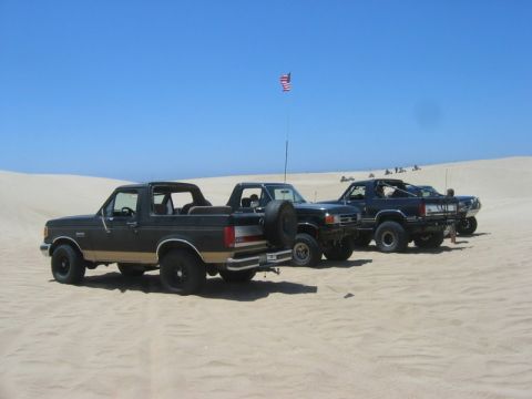 1989 Ford Bronco - Three Ford Broncos are parked side-by-side on sand dunes on a sunny day, with an American flag flying in the background.