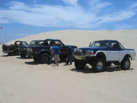 1989 Ford Bronco - Four trucks, including a 1989 Ford Bronco, are parked on sand dunes on a sunny day.