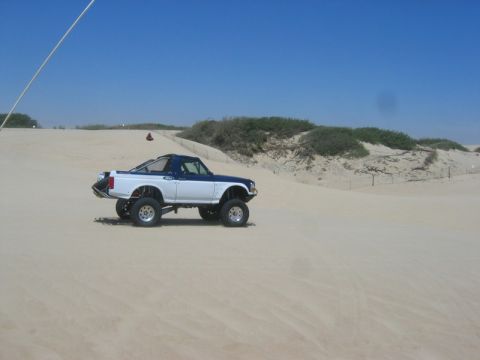 1989 Ford Bronco - A two-tone Ford Bronco is shown driving through a sandy landscape with dunes and sparse vegetation.