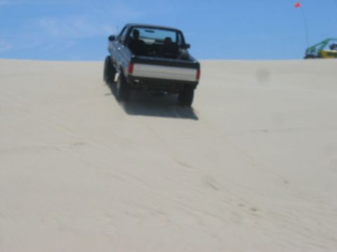 1989 Ford Bronco - A black pickup truck navigates a sandy incline under a clear blue sky, with another vehicle visible in the distance.