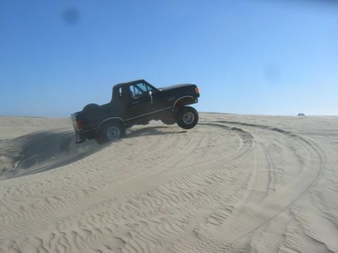 1989 Ford Bronco - A black 1989 Ford Bronco navigates sandy terrain, kicking up dust as it drives across the dunes.