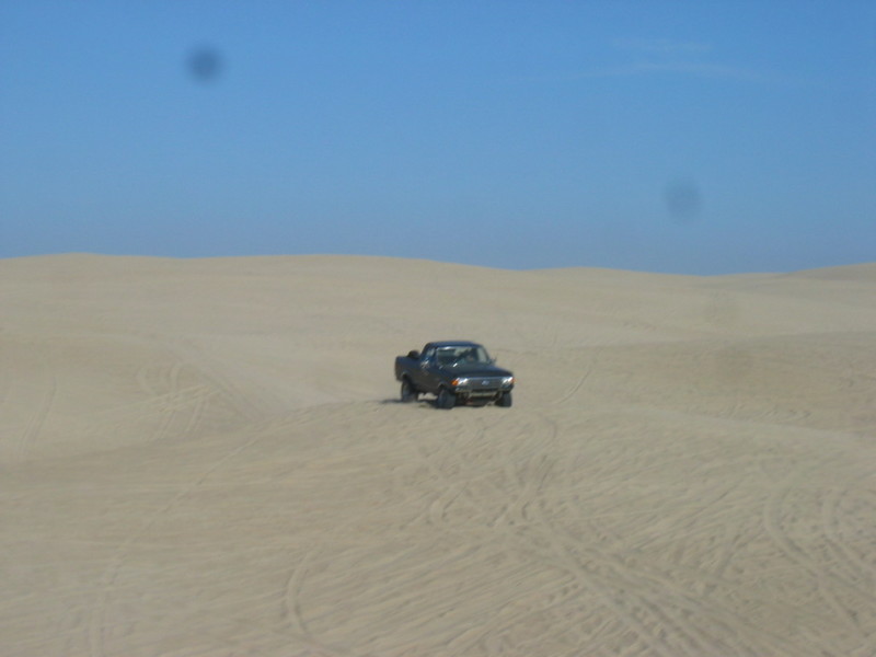 dark pickup truck driving on sand dunes under a clear blue sky