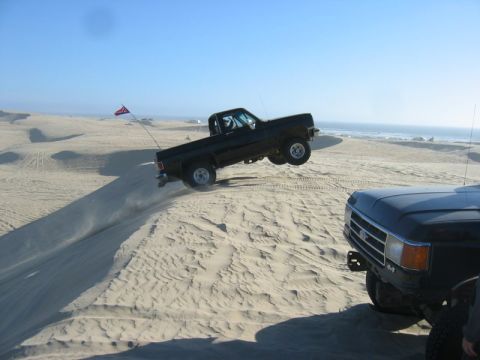 1989 Ford Bronco - A black pickup truck is captured mid-air as it jumps over a sand dune, with another truck visible in the foreground.