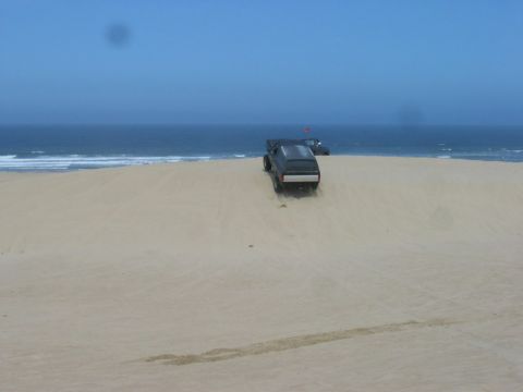 1989 Ford Bronco - Two SUVs navigate sandy terrain with the ocean in the distance under a clear blue sky.