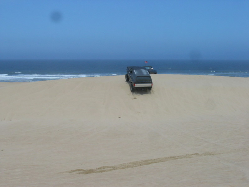 Black SUV driving up a sand dune, with another SUV behind it. Ocean and blue sky in the background