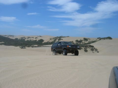 1989 Ford Bronco - A black 1987 Ford Bronco navigates a sandy terrain under a partly cloudy sky.