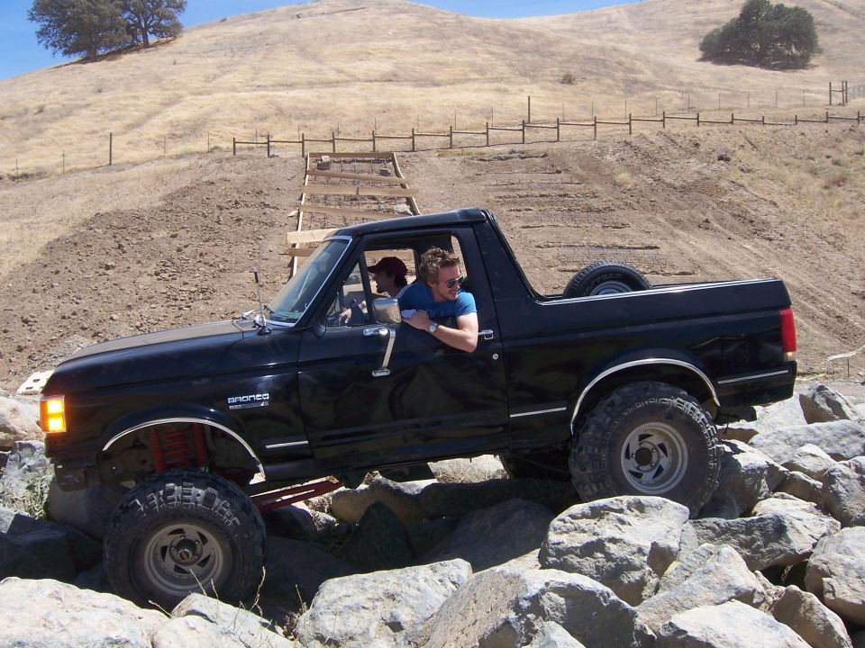 Black 1989 Ford Bronco with two people inside driving over large rocks on a dirt hill