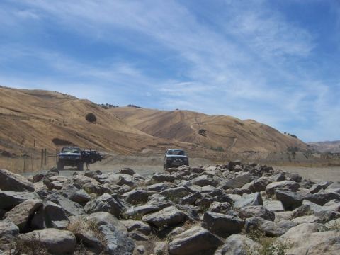 1989 Ford Bronco - Two SUVs navigate a rocky dirt road in a dry, hilly landscape under a partly cloudy sky.
