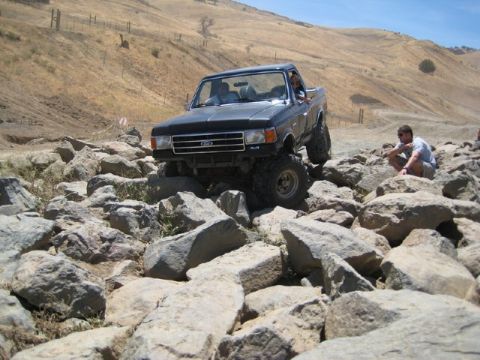 1989 Ford Bronco - A modified black Ford F-Series truck tackles a challenging rocky obstacle course on a sunny day.