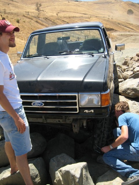 1989 Ford Bronco - A man inspects the front tire of a dark Ford truck navigating a rocky obstacle.